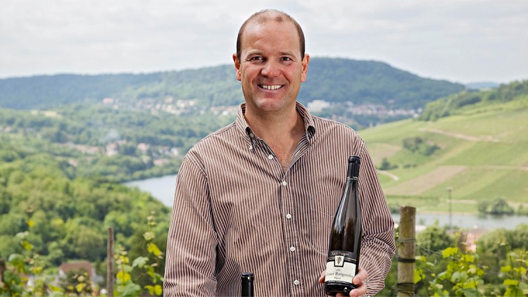 Person hält eine Flasche Grauer Burgunder mit Etikett „Karl Pegau“ in den Händen, im Hintergrund grüne Weinberge.