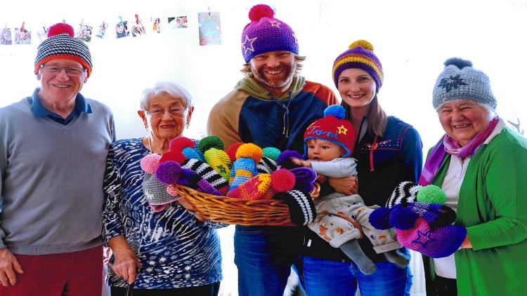 Gruppe von Menschen mit bunten Strickmützen hält einen gefüllten Korb mit weiteren farbenfrohen Mützen vor einem hellen Hintergrund.
