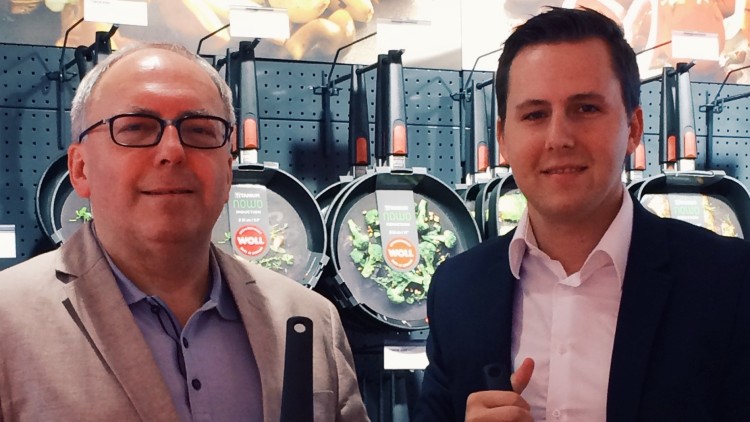 Zwei Personen halten WOLL-Pfannen vor einer Wand mit weiteren Pfannen und Koch-Zubehör.