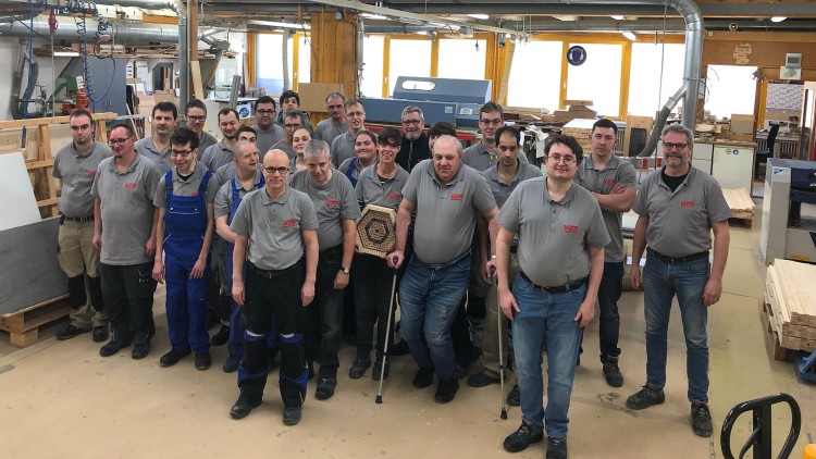 Gruppe von Menschen in Arbeitskleidung mit Holzplakette in einer Werkstatt, umgeben von Maschinen und Holzmaterialien.