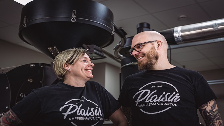 Zwei Personen tragen schwarze T-Shirts mit dem weißen Schriftzug „Plaisir Kaffeemanufaktur“ vor einer Kaffeeröstmaschine.
