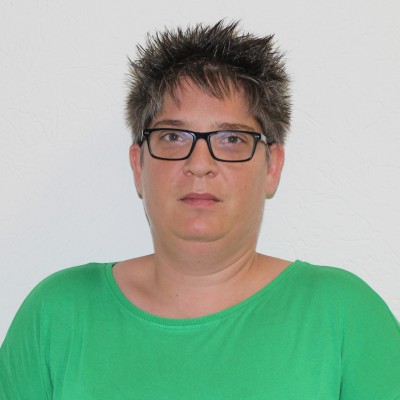 Person mit kurzen, dunkelbraunen und spitz nach oben gestylten Haaren trägt ein grünes Oberteil vor weißem Hintergrund.