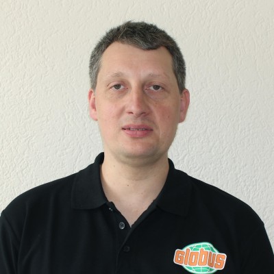 Person in schwarzem Poloshirt mit GLOBUS-Logo steht vor einer weißen, strukturierten Wand.