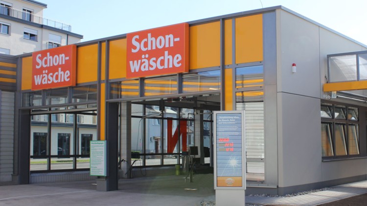 Ein Gebäude mit der Beschriftung „Schonwäsche“ in gelb-orange, links ein Schild mit Waschoptionen, rechts ein Baum.