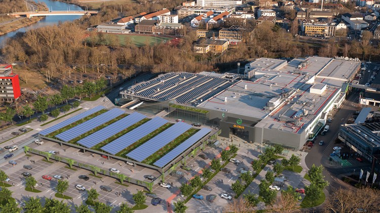 Luftaufnahme eines GLOBUS Marktes mit Parkplatz, Solarzellen auf Parkdach und Gebäudedach, umgeben von Bäumen und Stadtgebiet.