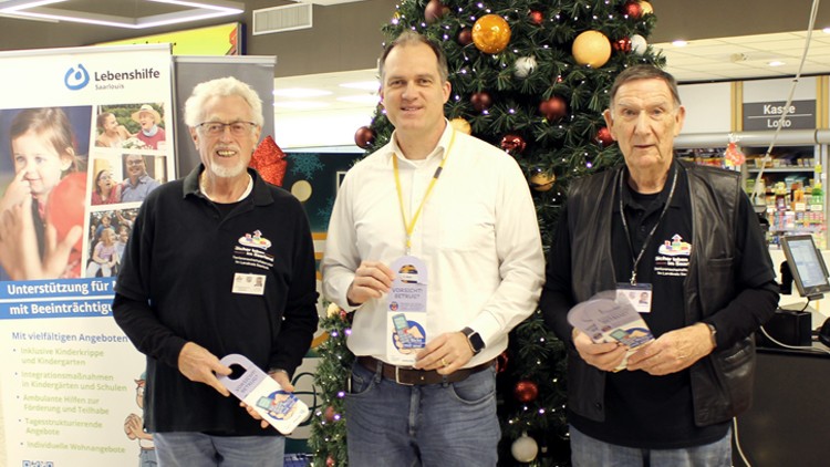 Drei Personen stehen vor einem dekorierten Weihnachtsbaum, halten Flyer der Lebenshilfe Saarlouis und stehen neben einem Infostand.