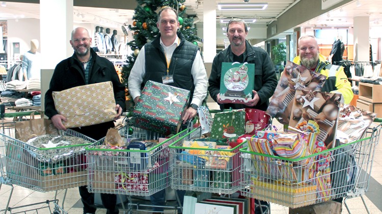 Sechs Einkaufswagen voller Geschenke vor einem geschmückten Weihnachtsbaum in einem Laden mit Kleidung im Hintergrund.