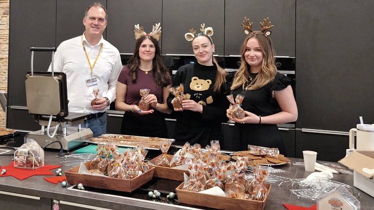 Personen stehen hinter einem Tisch mit verpackten Lebkuchen, auf dem eine Waffeleisen-Maschine und Dekorationen platziert sind.