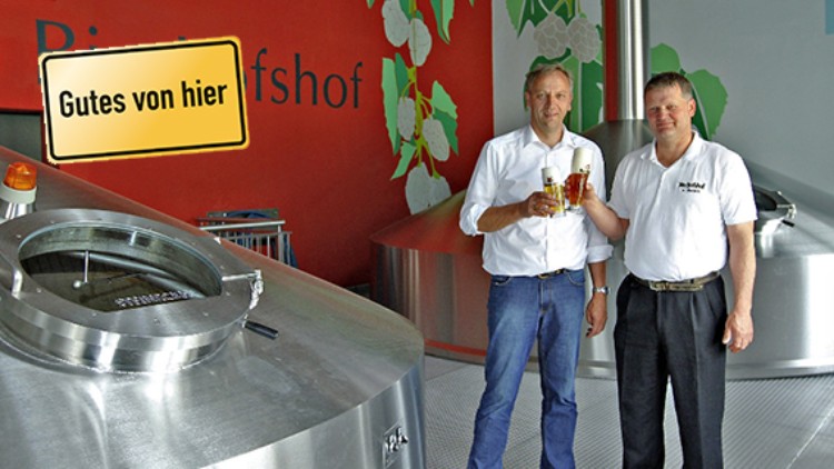 Zwei Personen halten je ein Bierglas vor Gärbehältern, an der Wand steht „Gutes von hier“ und „Brauhof“.