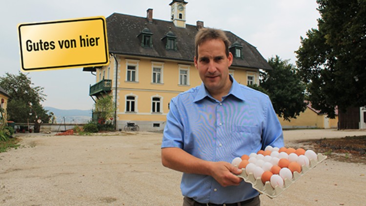 Mann in blauem Hemd hält eine Schale mit braunen und weißen Eiern vor einem gelben Gebäude, Schild mit „Gutes von hier“.