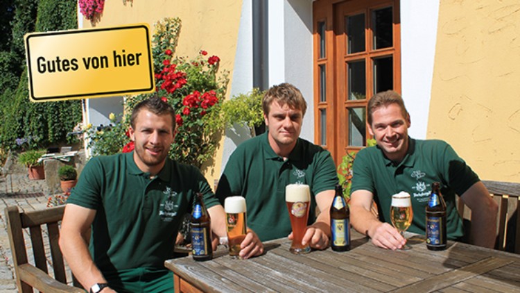 Drei Personen in grünen Poloshirts sitzen an einem Holztisch im Freien mit verschiedenen Bierflaschen und -gläsern, Schild „Gutes von hier“.