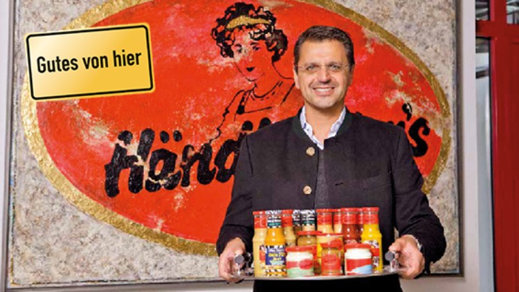Mann in dunkler Kleidung hält ein Tablett mit verschiedenen Gewürzgläsern vor einem großen roten Händlmaier-Logo, Schild \