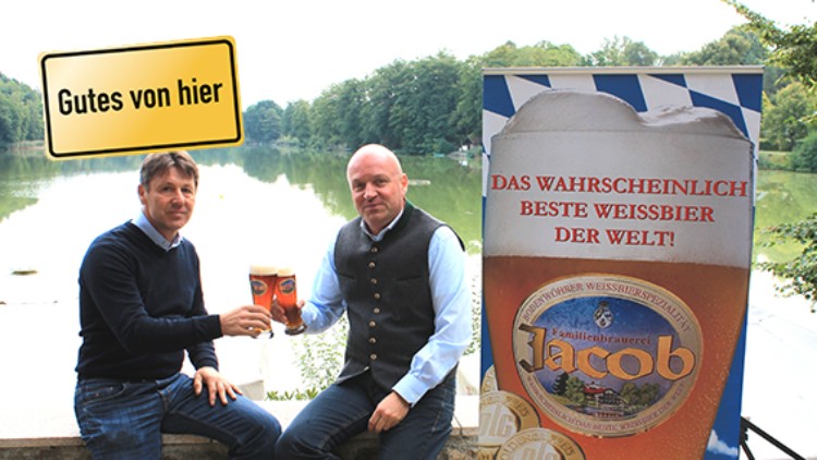 Zwei Personen stoßen mit Weißbier an, daneben ein Plakat mit „Das wahrscheinlich beste Weißbier der Welt!“ und „Gutes von hier“.