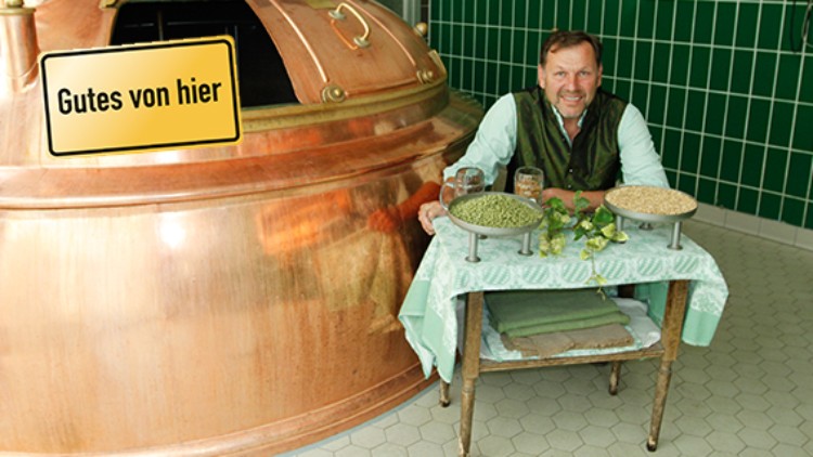 Eine kupferne Brauanlage neben einem Tisch mit Gläsern, Getreideschalen und der Aufschrift „Gutes von hier“.