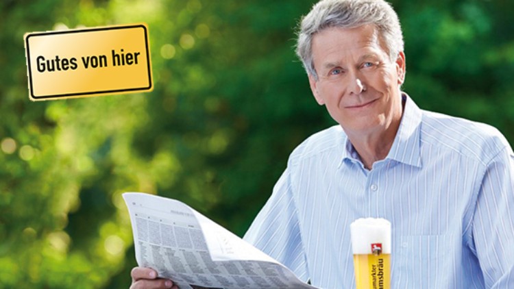 Person in hellem Hemd hält eine Zeitung, vor ihr steht ein Bierglas, links oben steht „Gutes von hier“ auf gelbem Schild.