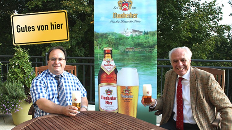 Zwei Personen sitzen an einem Holztisch, halten Biergläser und eine Werbetafel für Naabecker Bier steht im Hintergrund.
