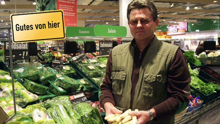 Person hält Spargelbündel vor einer Auswahl an grünem Salat mit dem Schild „Gutes von hier“ im Hintergrund.