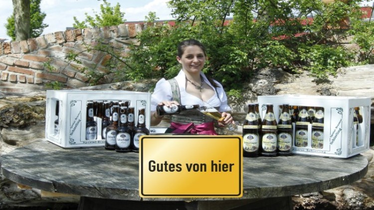 Zwei Getränkekisten und eine Frau in Tracht mit einer Bierflasche, davor ein gelbes Schild mit der Aufschrift Gutes von hier.