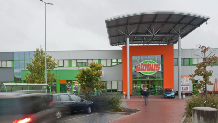 Eingangsbereich eines GLOBUS Marktes mit orangener Fassade, grünem Logo, Parkplätzen und einem Überdach mit Stahlträgern.