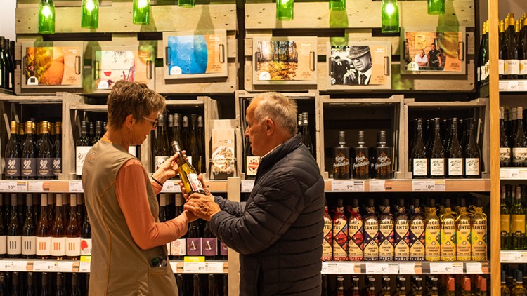 Zwei Personen stehen vor einem Weinregal in einer WEINLOUNGE, eine Person hält eine Weinflasche in der Hand.