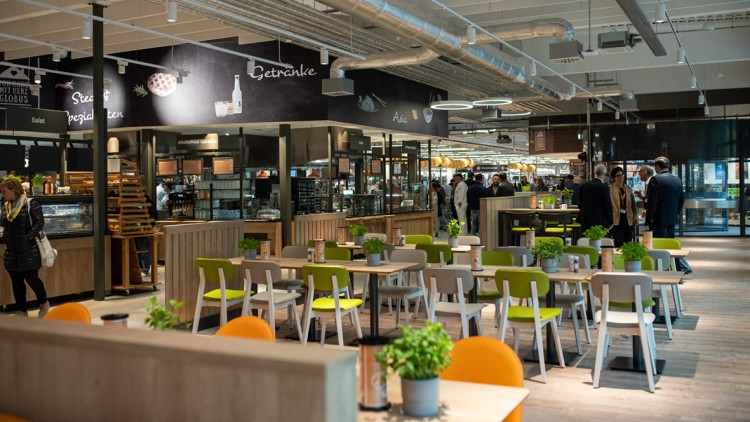 Offene Gastronomiefläche mit Tischen, grünen und orangen Stühlen, Pflanzen und Rückbuffet mit Schriftzügen wie „Getränke“.