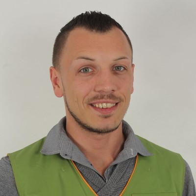 Person in grauem Hemd und grüner Weste mit orangefarbener Umrandung vor neutralem Hintergrund.