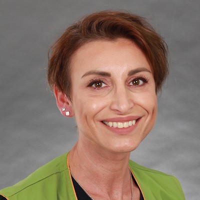 Person trägt grüne Arbeitskleidung mit orangefarbenen Nähten und silberne quadratische Ohrringe, grauer Hintergrund.