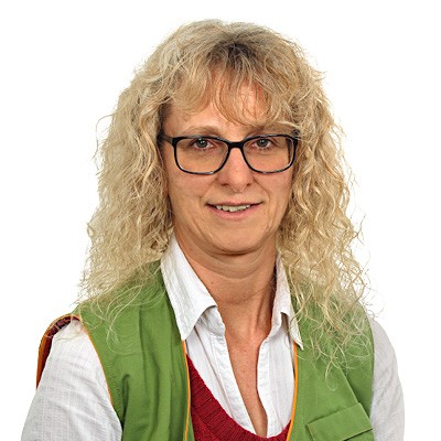 Person mit blonden, lockigen Haaren trägt weiße Bluse, roten Pullover und grüne Weste vor weißem Hintergrund.