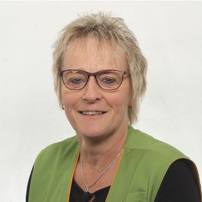 Person mit kurzem, blondem Haar trägt eine grüne Weste mit orangefarbener Paspelierung und eine silberne Kette.