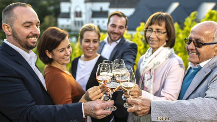 Mehrere Personen stoßen mit gefüllten Weingläsern vor einem weißen Gebäude und Weinreben im Hintergrund an.