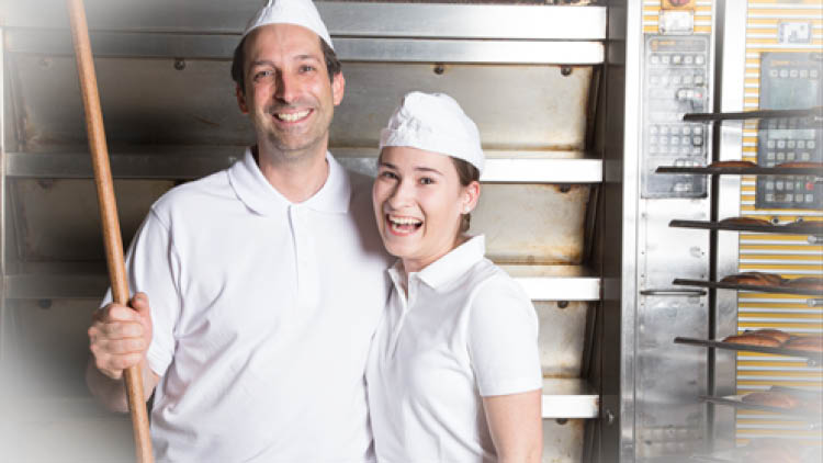 Zwei Personen in weißer Arbeitskleidung stehen vor einem Backofen, eine hält einen Holzbackschieber.