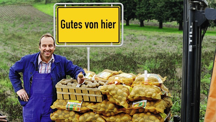 Stapelnetze mit Kartoffeln und eine Holzkiste stehen vor einer Wiese, daneben ein gelbes Schild mit der Aufschrift Gutes von hier.
