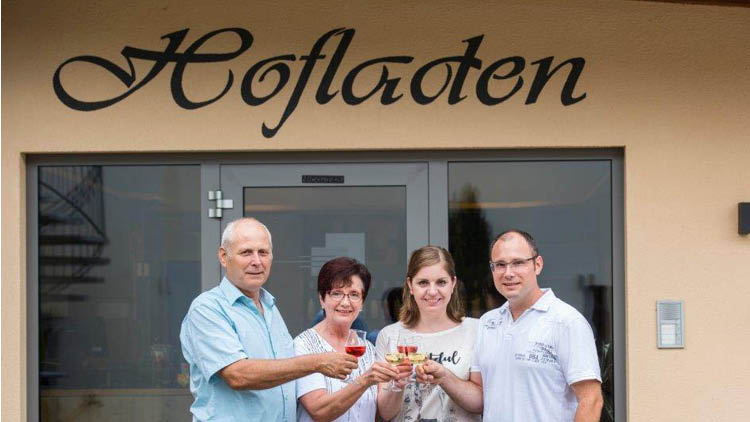 Vier Personen stoßen mit gefüllten Weingläsern vor dem Schriftzug „Hofladen“ und einer Glastür an.