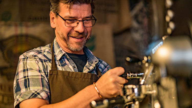 Person mit kariertem Hemd und brauner Schürze bedient eine silberne Kaffeemaschine in einer warm beleuchteten Umgebung.