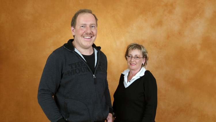 Person in dunkler Jacke und Person in dunklem Pullover mit weißem Kragen vor einem orangefarbenen, strukturierten Hintergrund.