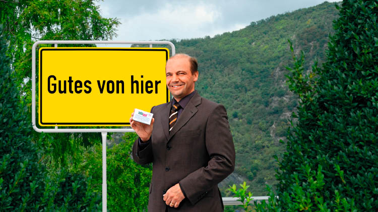 Person in Anzug hält eine Packung Sebamed-Creme vor einem gelben Schild mit der Aufschrift Gutes von hier, grüne Landschaft im Hintergrund.