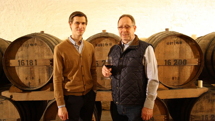 Zwei Personen stehen vor mehreren Holzfässern in einem Weinkeller, eine Person hält ein Glas Rotwein in der Hand.