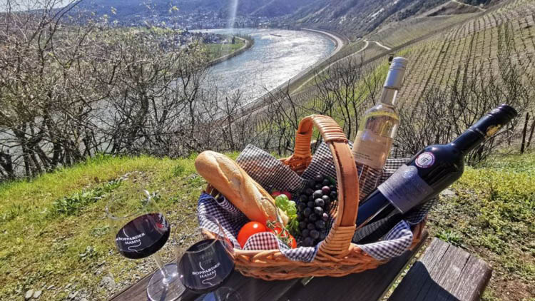 Geflochtener Picknickkorb mit Baguette, Trauben, Tomaten und zwei Weinflaschen auf Holztisch, Fluss und Weinberge im Hintergrund.