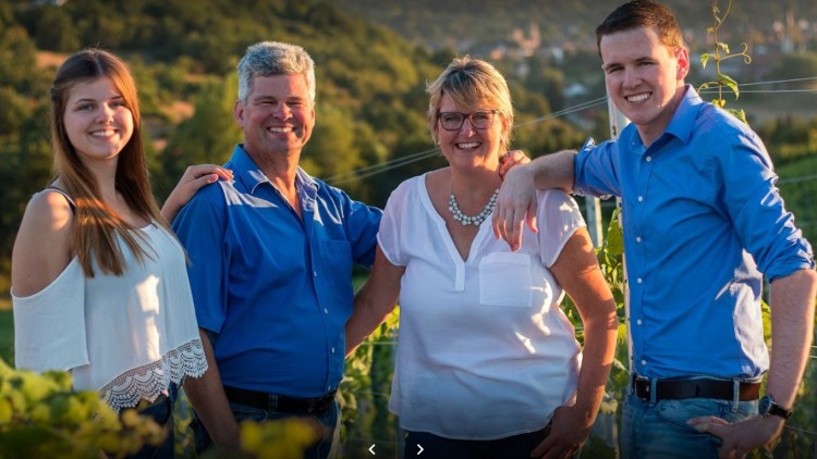 Vier Personen stehen lächelnd in einem Weinberg, umgeben von grünen Reben und einer hügeligen Landschaft im Hintergrund.