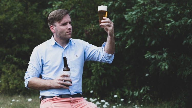 Person in blauem Hemd und rosafarbenen Hosen hält ein Bierglas hoch, in der anderen Hand eine Bierflasche, im Hintergrund Bäume.