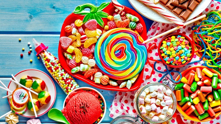 Verschiedene bunte Süßigkeiten wie Lutscher, Schokolade, Gummibärchen und Marshmallows sind auf einem blauen Holztisch arrangiert.