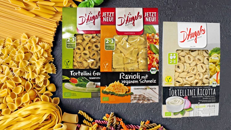 Drei Packungen D'Angelo Pasta-Sorten, umgeben von roher Pasta in verschiedenen Formen auf grauem Hintergrund.