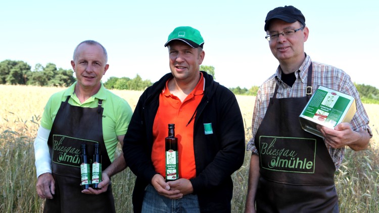 Drei Personen tragen Schürzen mit der Aufschrift „Bliesgau Ölmühle“ und halten Produkte wie Ölflaschen und eine Verpackung.