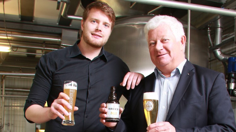 Zwei Personen in dunkler Kleidung halten Biergläser und eine Bierflasche mit der Aufschrift „BRUCH No1“ in einer Brauerei.