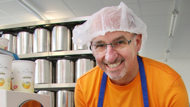 Person in orange Kleidung mit blauem Schürzenband, trägt Haarnetz, im Hintergrund Metallbehälter und Eisbecher mit Aufschrift Pistazie.
