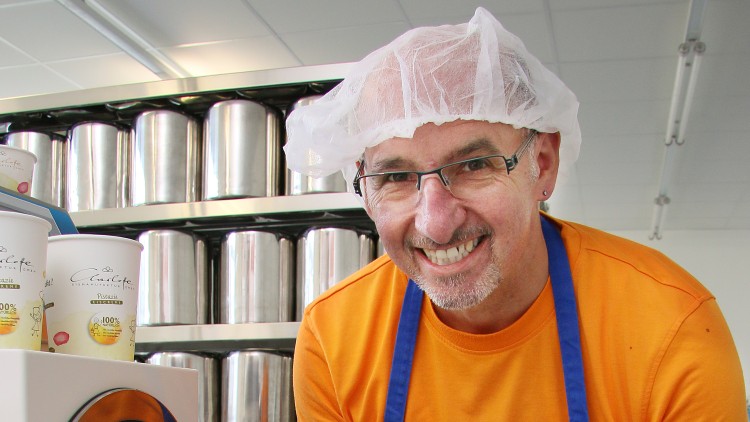 Person in orange Oberteil mit blauer Schürze und Haarnetz vor Regalen mit silbernen Behältern und Eiscups auf einem Aufsteller.