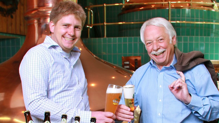 Zwei Personen stoßen mit Gläsern gefüllten Bier an, im Hintergrund eine Brauereikulisse mit Kupferkesseln und grünen Fliesen.