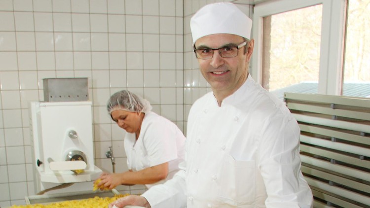 Zwei Personen in weißer Arbeitskleidung arbeiten in einer Küche mit gefliesten Wänden und großen Mengen gelber Teigwaren.
