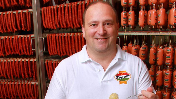 Person in weißem Polo mit „Kunzler“-Logo vor Regalen mit geräucherten Würsten, hält eine Gabel mit Sauerkraut.