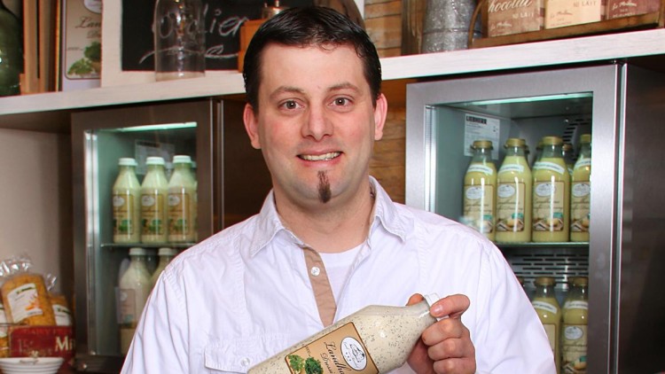 Person in weißem Hemd hält eine Flasche mit cremigem Dressing vor Kühlregalen mit weiteren Flaschen desselben Produkts.